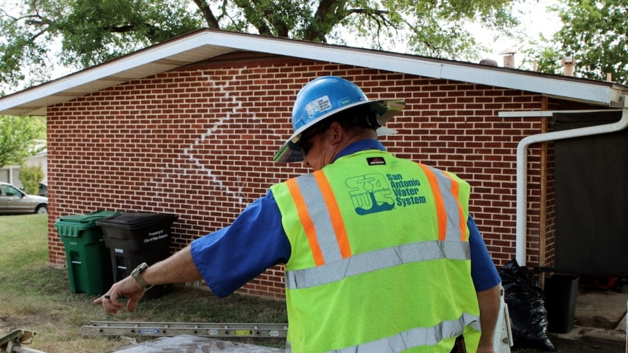 Utility worker in safety gear inspects site near brick building and trash bins on a sunny day